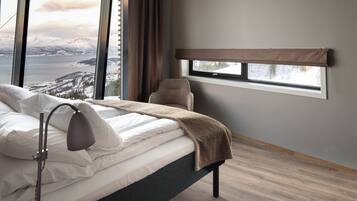A bedroom with a bed, a bedside lamp, a window with a view of mountains and a body of water, and a chair.