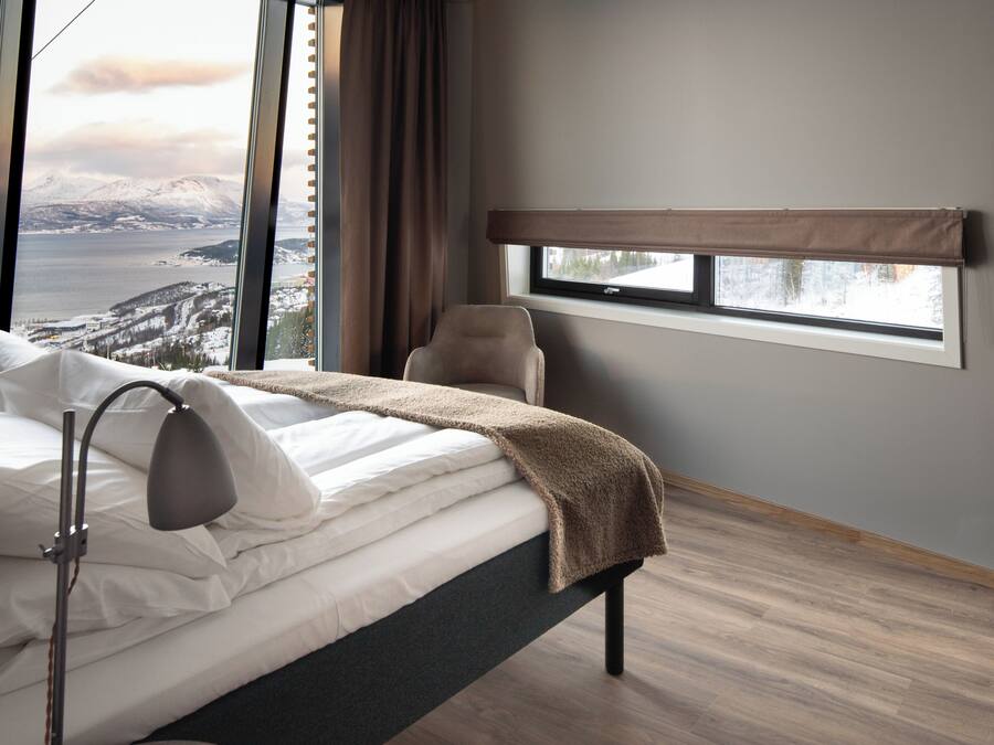 A bedroom with a bed, a bedside lamp, a window with a view of mountains and a body of water, and a chair.
