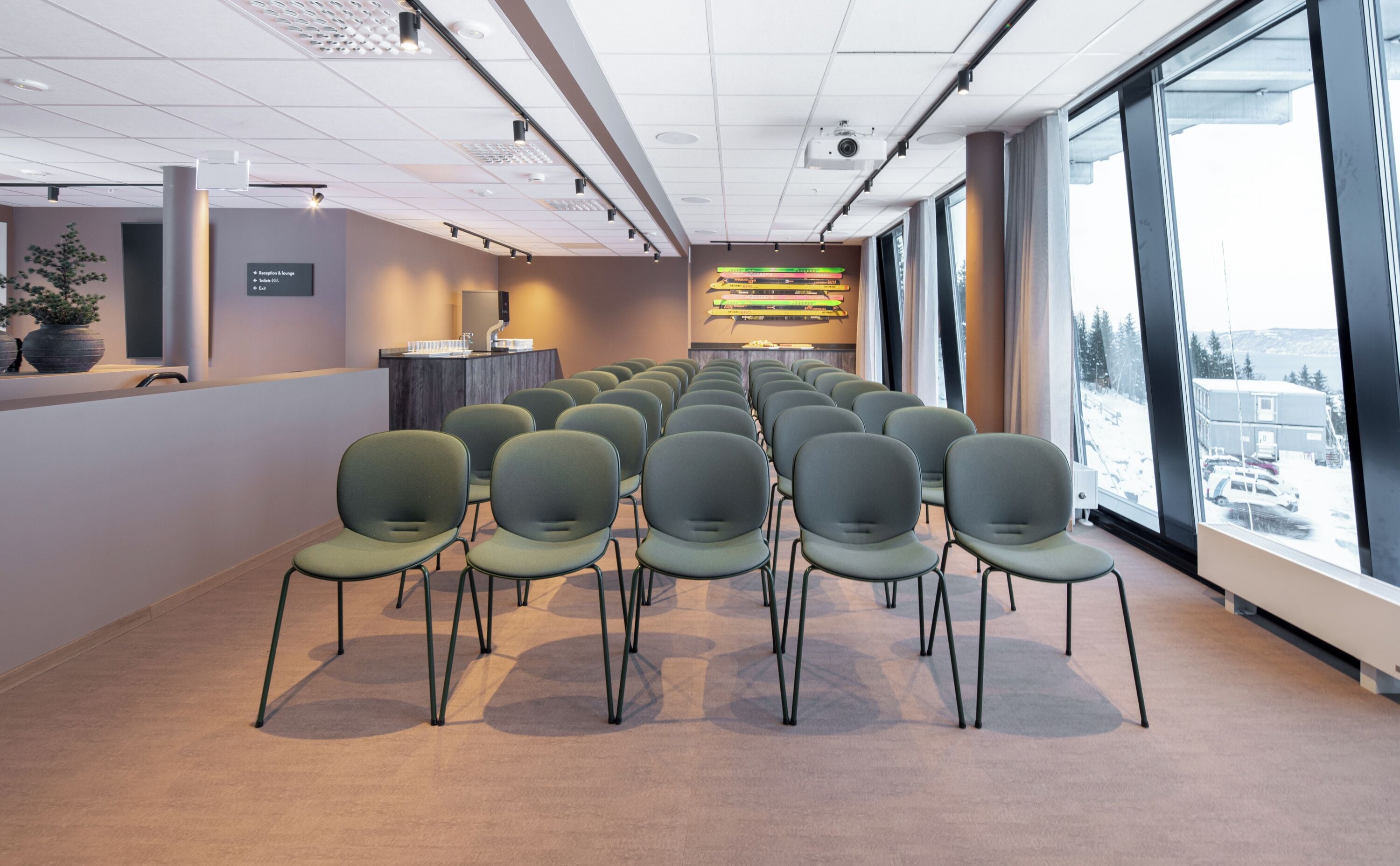 Un salón de conferencias con filas de sillas, una mesa de recepción y un amplio ventanal con vistas a un paisaje nevado.