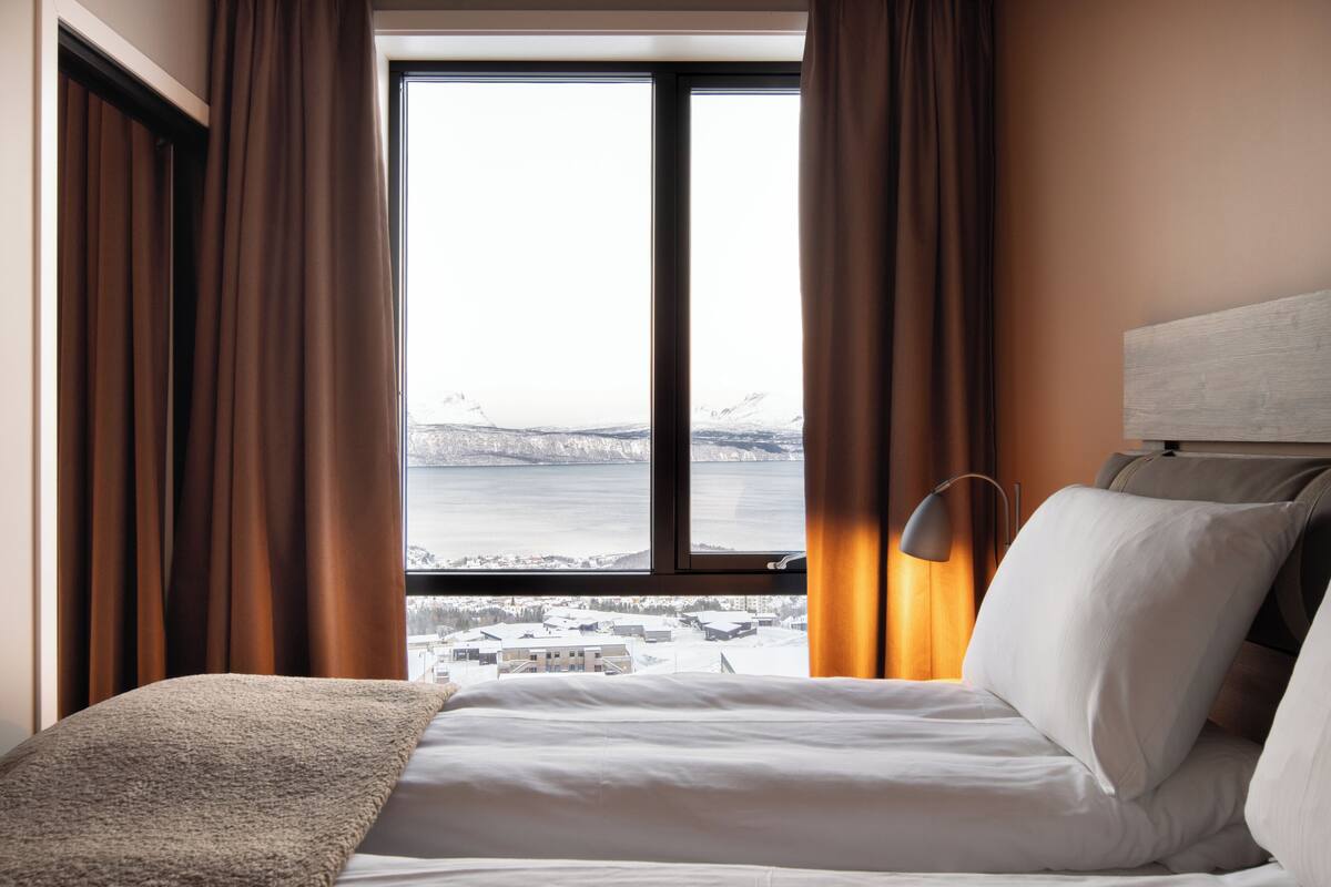 A hotel room with a bed, a view of a body of water and mountains, and a lamp.