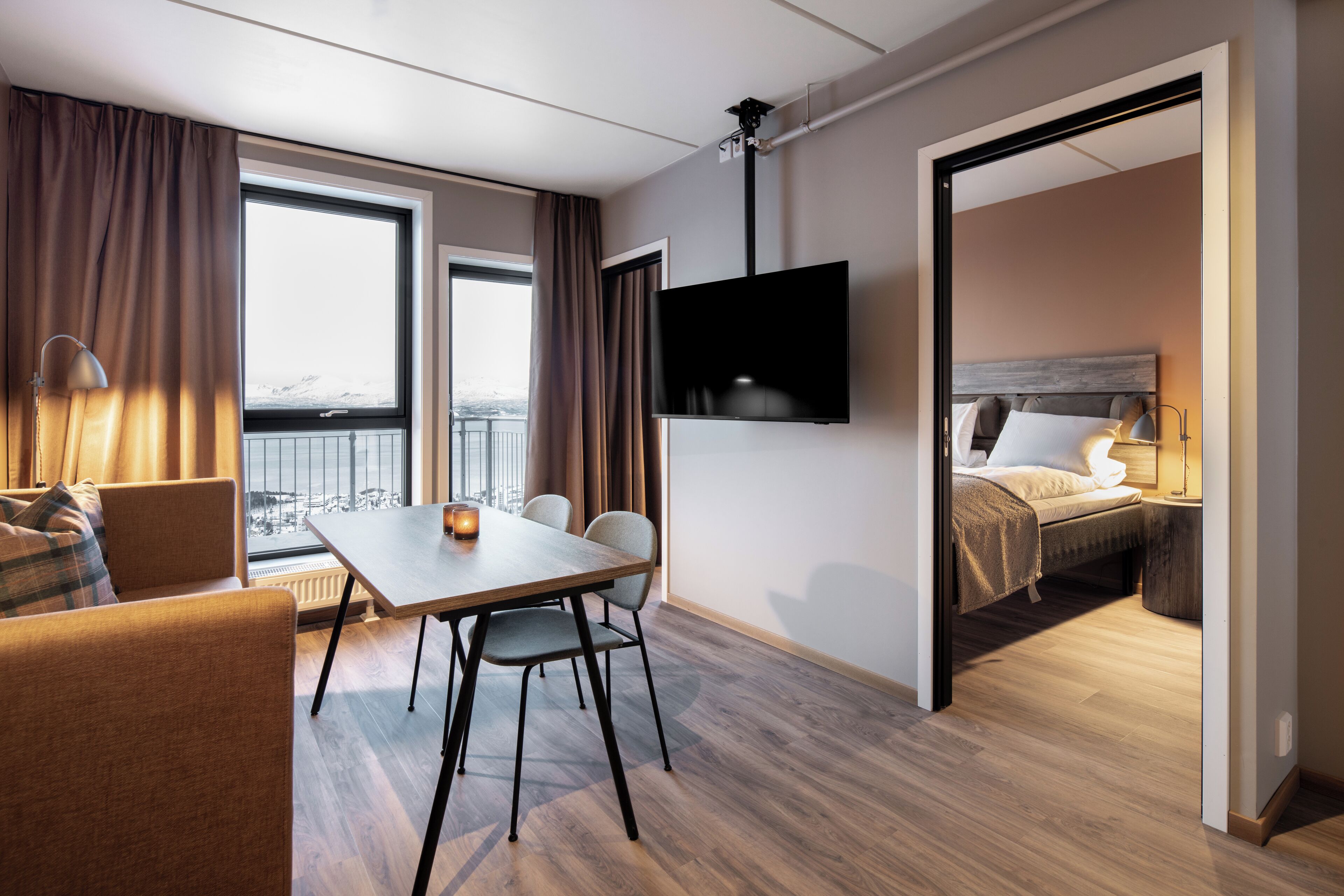 Habitación de hotel con zona de comedor, televisor montado en la pared, cama con cabecera y vistas a la ciudad a través de amplios ventanales.