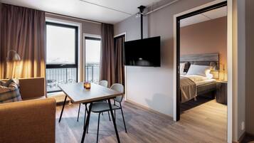 A hotel room with a dining area, a TV mounted on the wall, a bed with a headboard, and a view of the city through large windows.