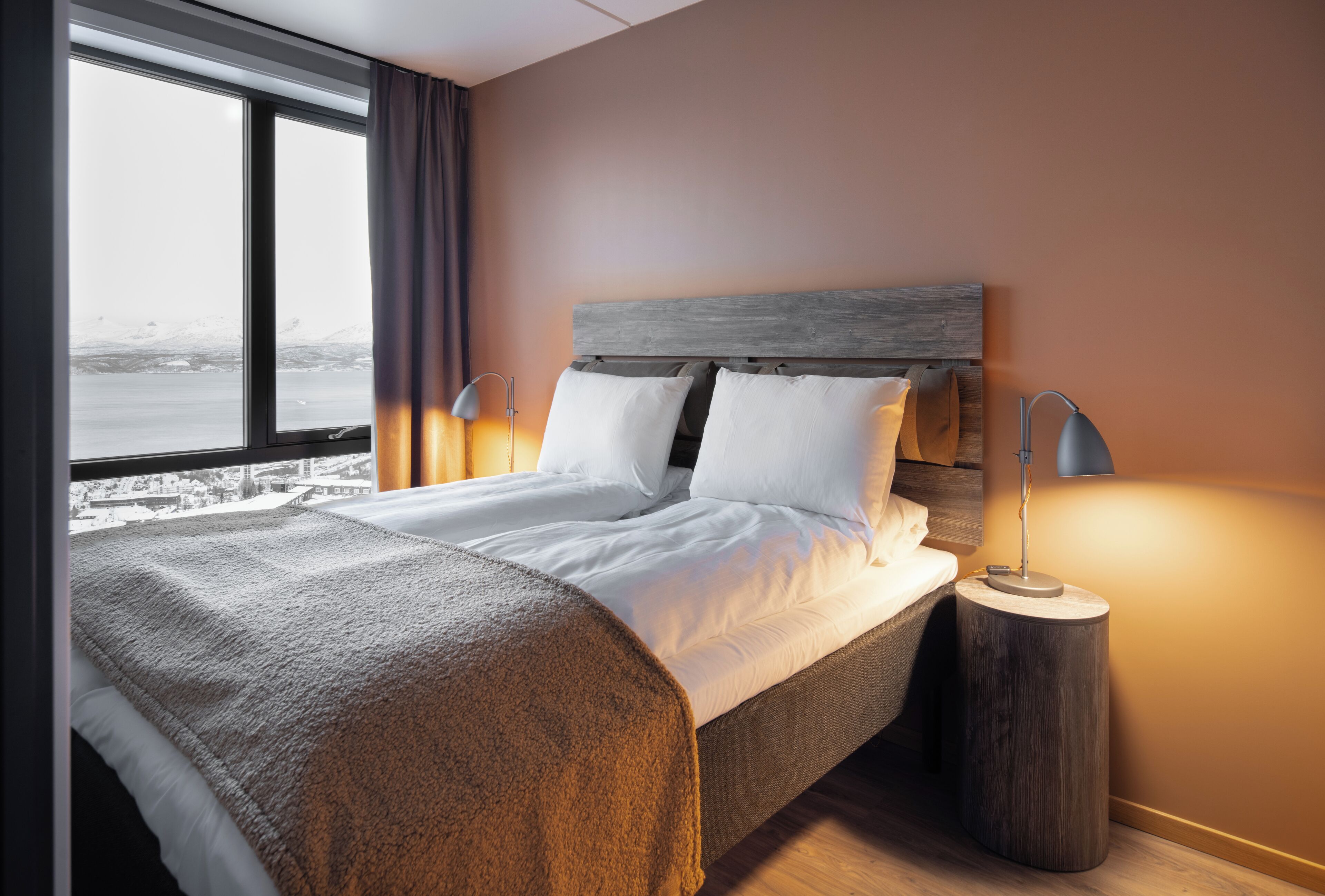 A neatly made bed with white linens and a brown throw blanket, a wooden headboard, and a bedside lamp on a round wooden nightstand.