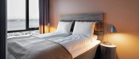 A neatly made bed with white linens and a brown throw blanket, a wooden headboard, and a bedside lamp on a round wooden nightstand.