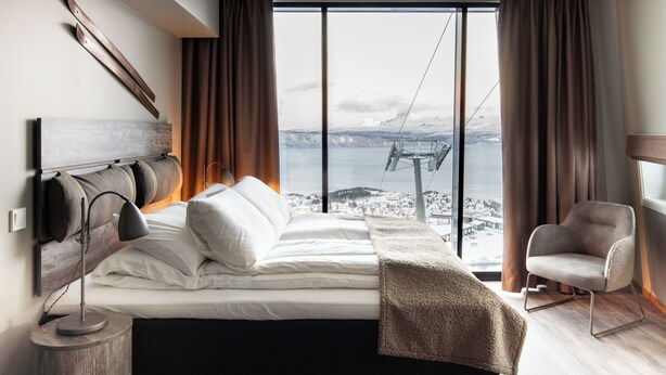 Une chambre moderne avec un grand lit, une table de chevet, une chaise et une vue sur un pont et le paysage urbain, le tout visible à travers une grande fenêtre.