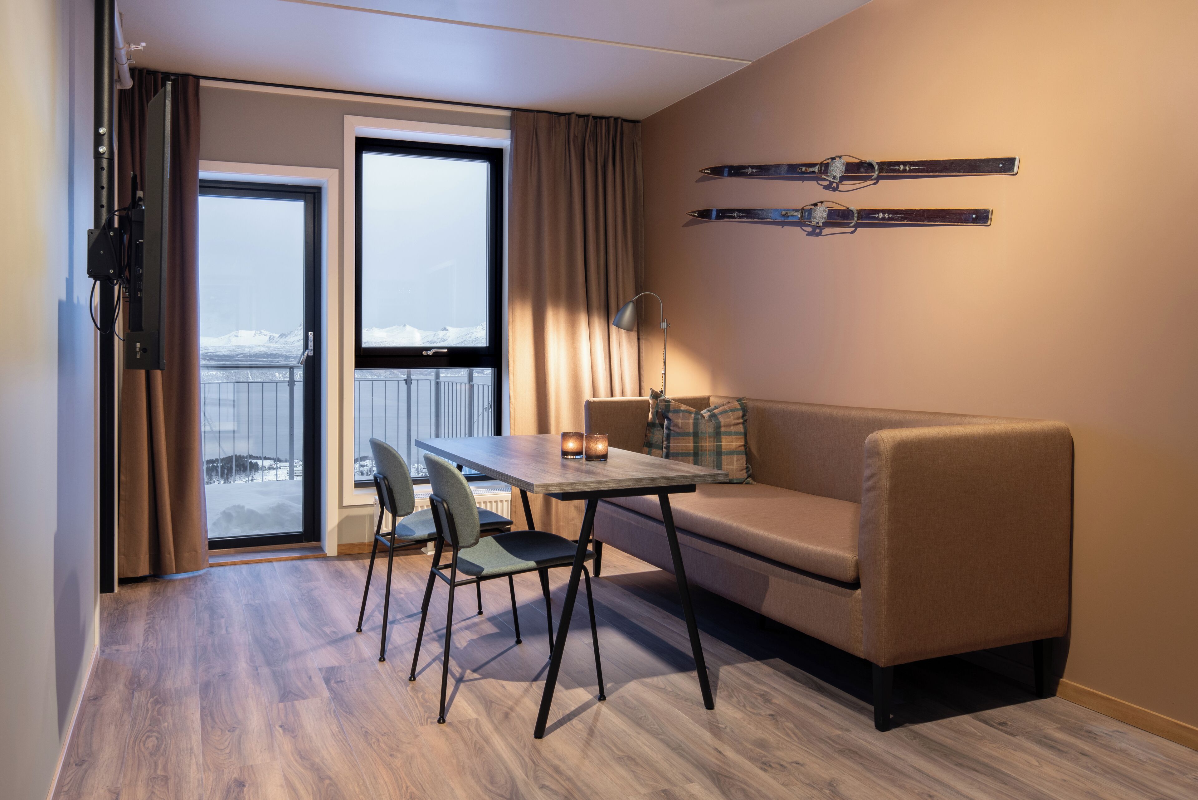 Ein Hotelzimmer mit Holzboden, einer Couch, einem Tisch mit Stühlen, einem Fenster mit Vorhängen und an der Wand befestigten Skiern.