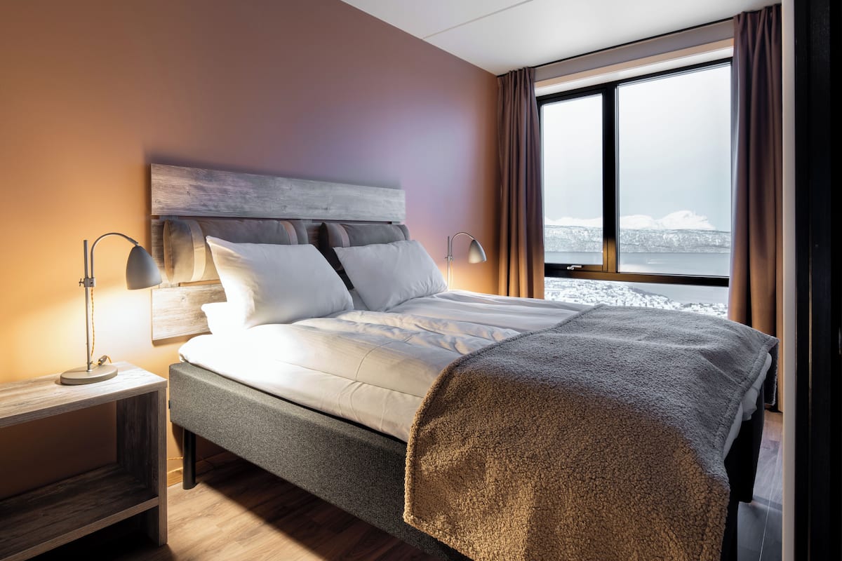 A modern hotel room with a large bed, a bedside table, a lamp, and a view of a snowy landscape through the window.