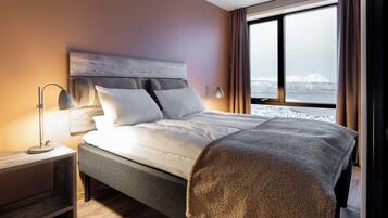 Ein modernes Hotelzimmer mit einem großen Bett, einem Nachttisch, einer Lampe und Blick auf eine verschneite Landschaft durch das Fenster.