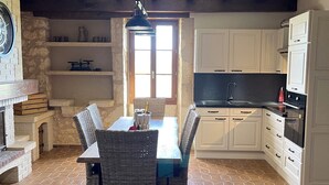 Un coin repas agrémenté d’une cheminée en briques, d’une table avec des chaises et d’une vue sur la cuisine, qui présente des armoires blanches et un comptoir noir.