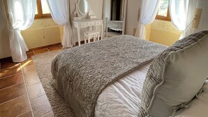 Une chambre avec un plafond en bois, un lit recouvert d’une couette à motifs, une table de chevet, un miroir et une fenêtre avec des rideaux.