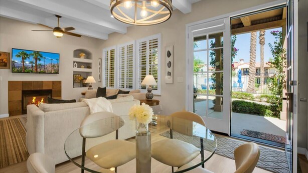 A modern living room with a glass coffee table, white sofas, a fireplace, and a view of the outdoor patio. - Poolside Paradise 3 | 3BR Legacy Villa (La Quinta)