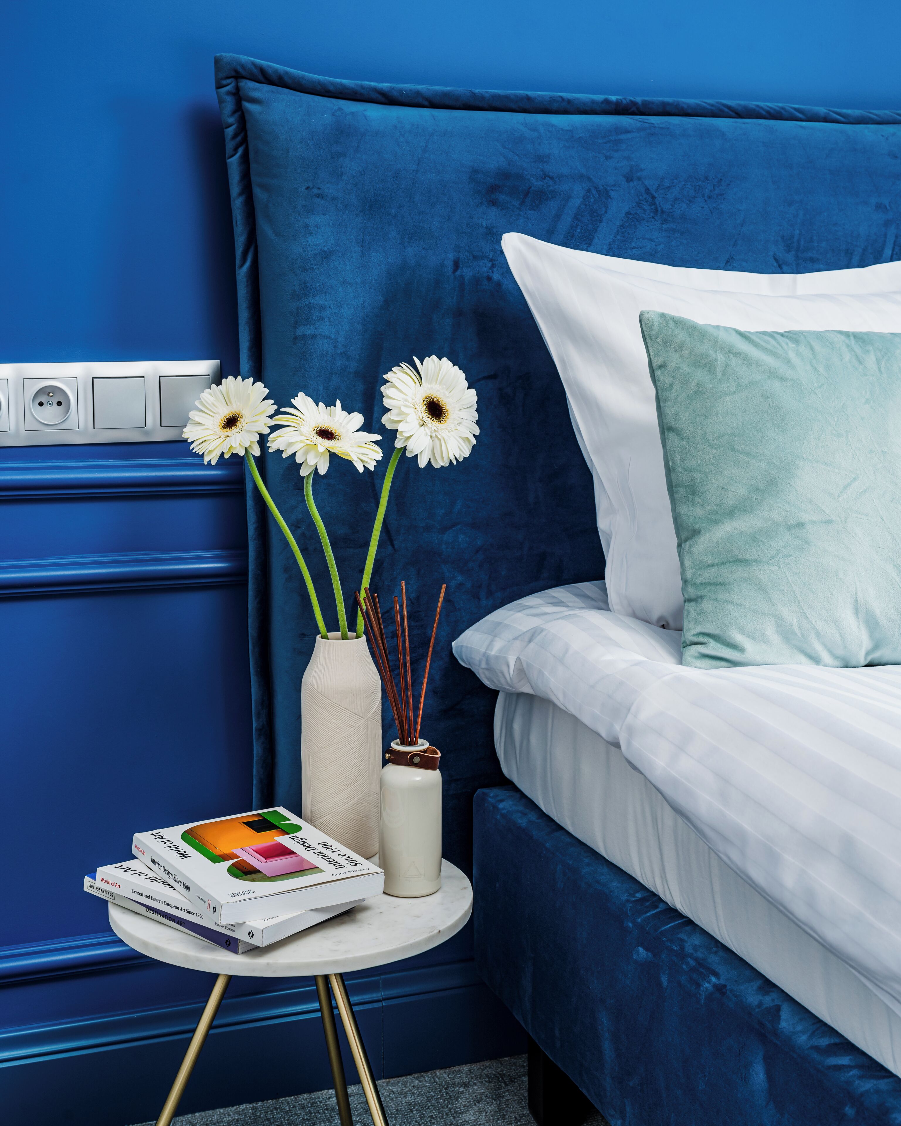 Un dormitorio con pared azul, una cama blanca con almohada turquesa, una mesita de noche blanca con libros y un florero con flores.