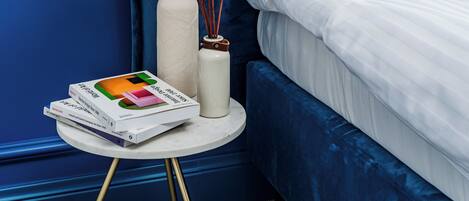 A bedroom with a blue wall, a white bed with a teal pillow, a white nightstand with books and a vase with flowers.