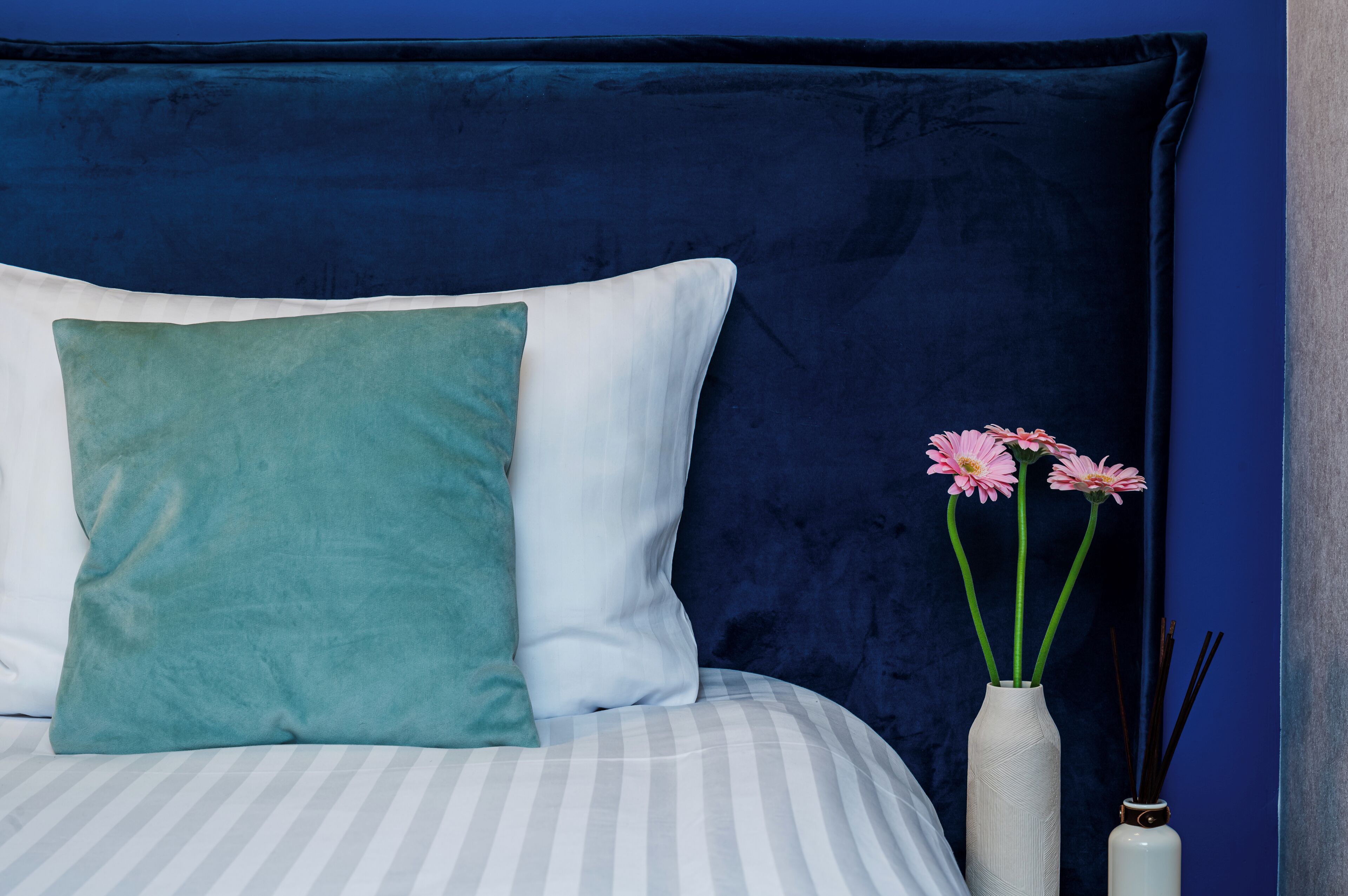 Una cama con un cojín azul verdoso y blanco, un florero blanco con flores rosadas y un florero blanco con ramas de caña.