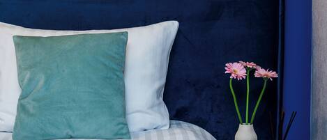 Una cama con un cojín azul verdoso y blanco, un florero blanco con flores rosadas y un florero blanco con ramas de caña.