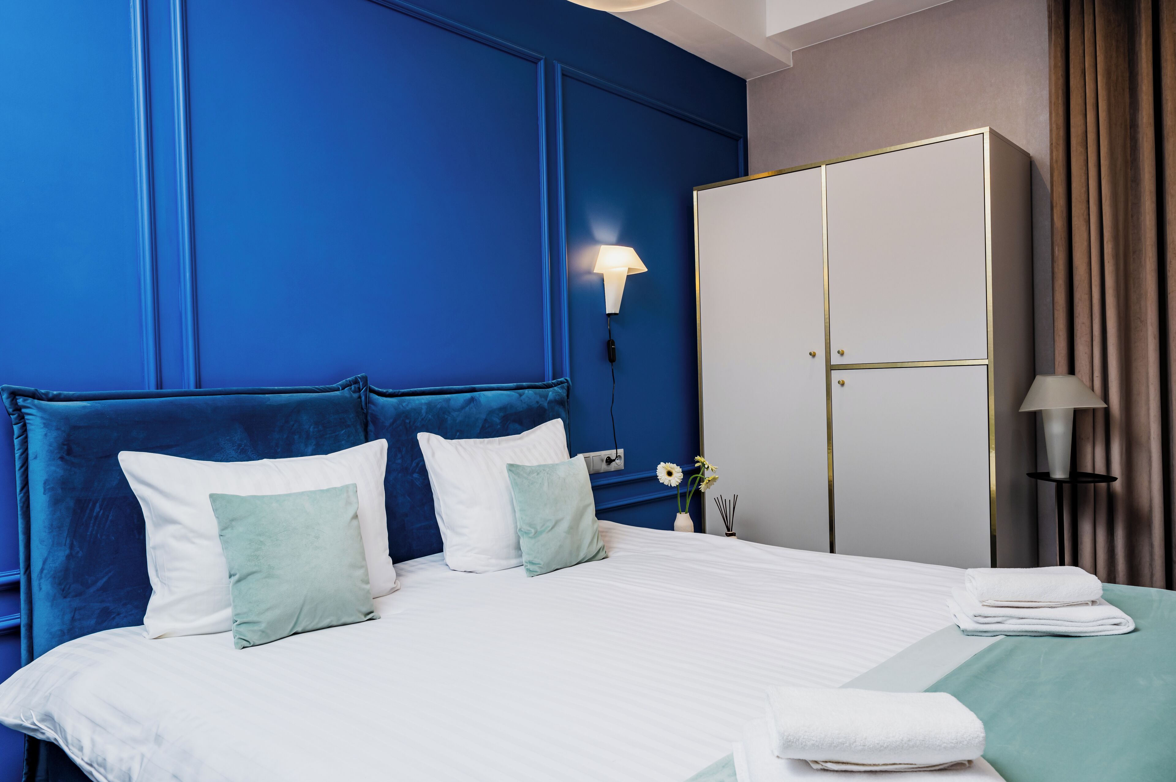 A neatly made bed with white and light blue pillows, a blue headboard, and a white wardrobe with gold accents.