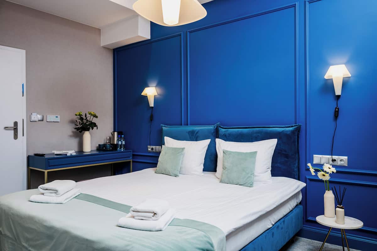 A modern hotel room with a large bed, bedside lamps, a small table with a vase of flowers, and a desk with a coffee machine.