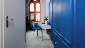 Un pasillo con una puerta azul, un escritorio blanco con un florero y una silla azul, además de una escalera con una puerta de madera.
