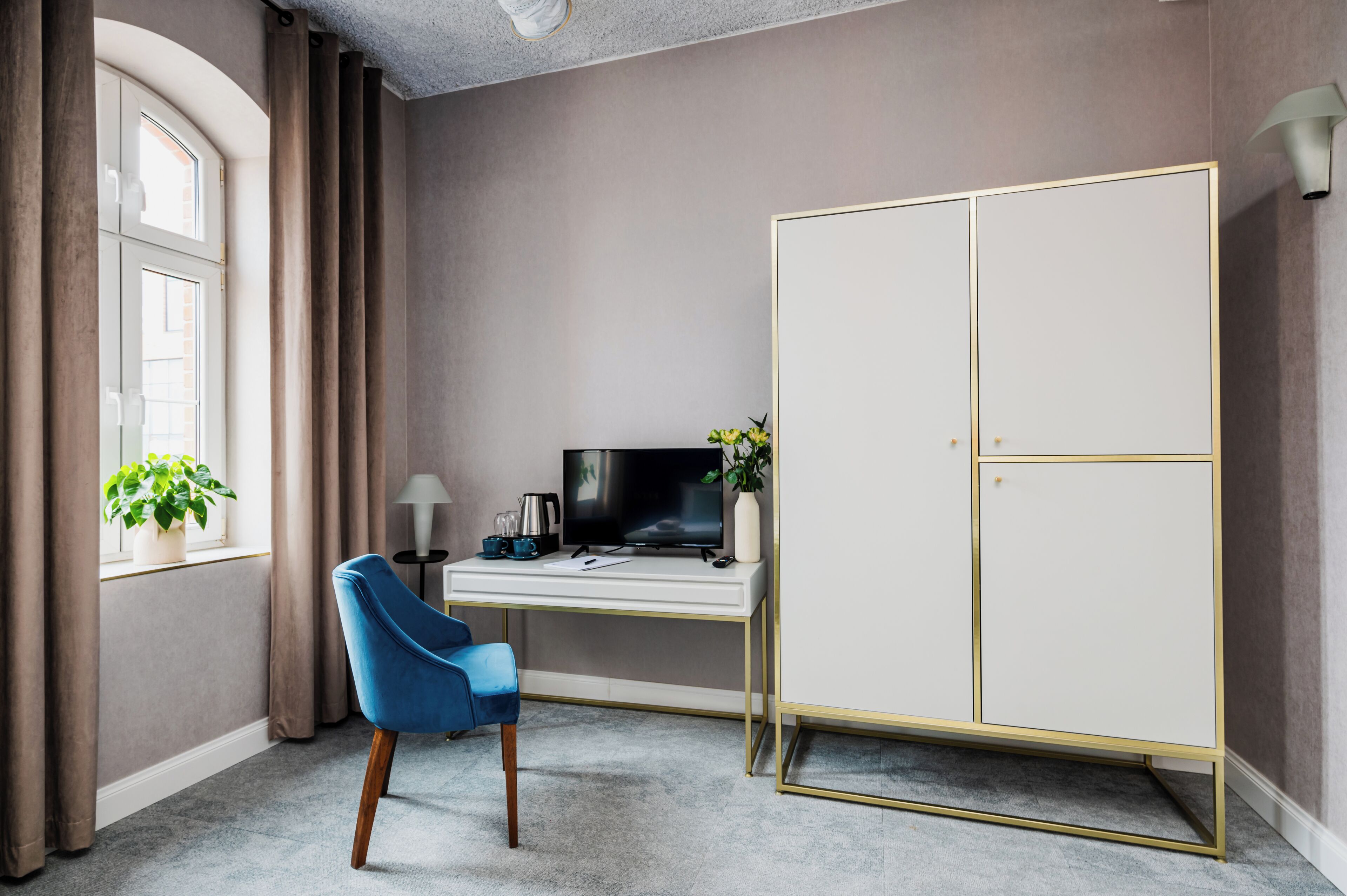 A room with a desk, a television, a wardrobe, a blue chair, and a window with curtains.