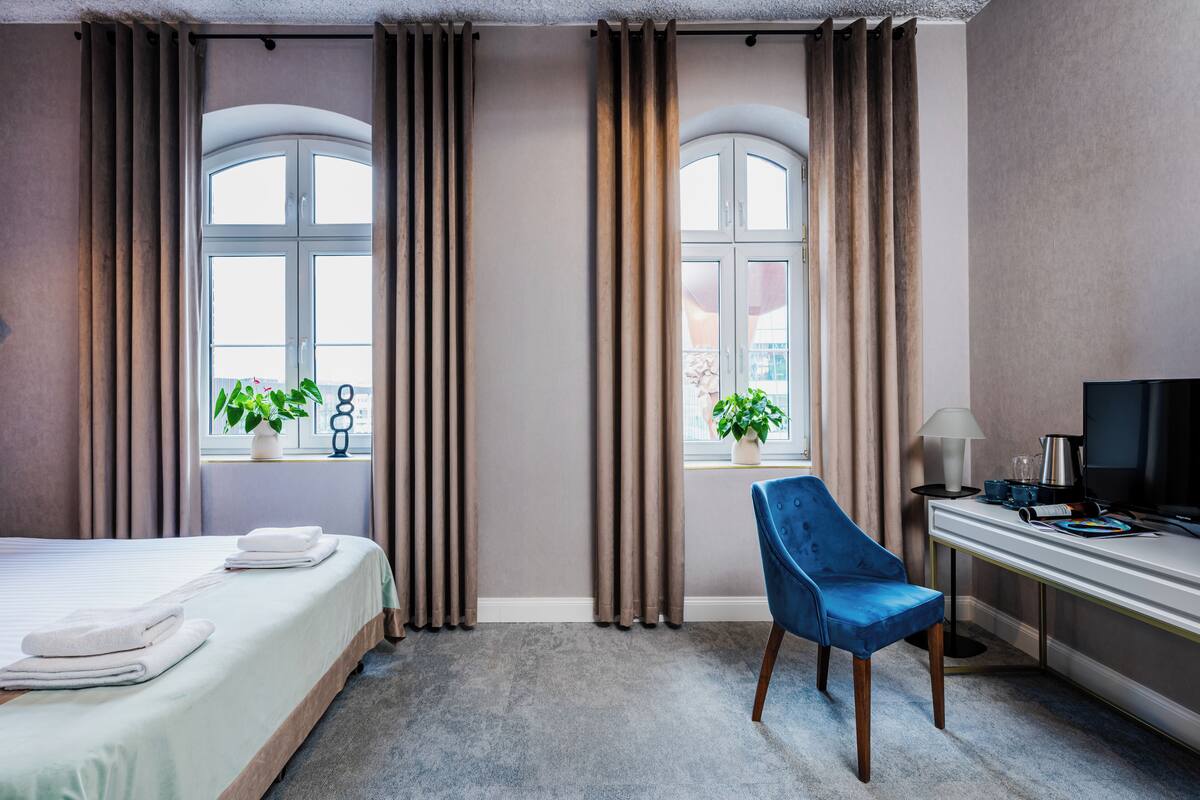 A hotel room with a bed, a desk, a chair, and two windows with curtains.
