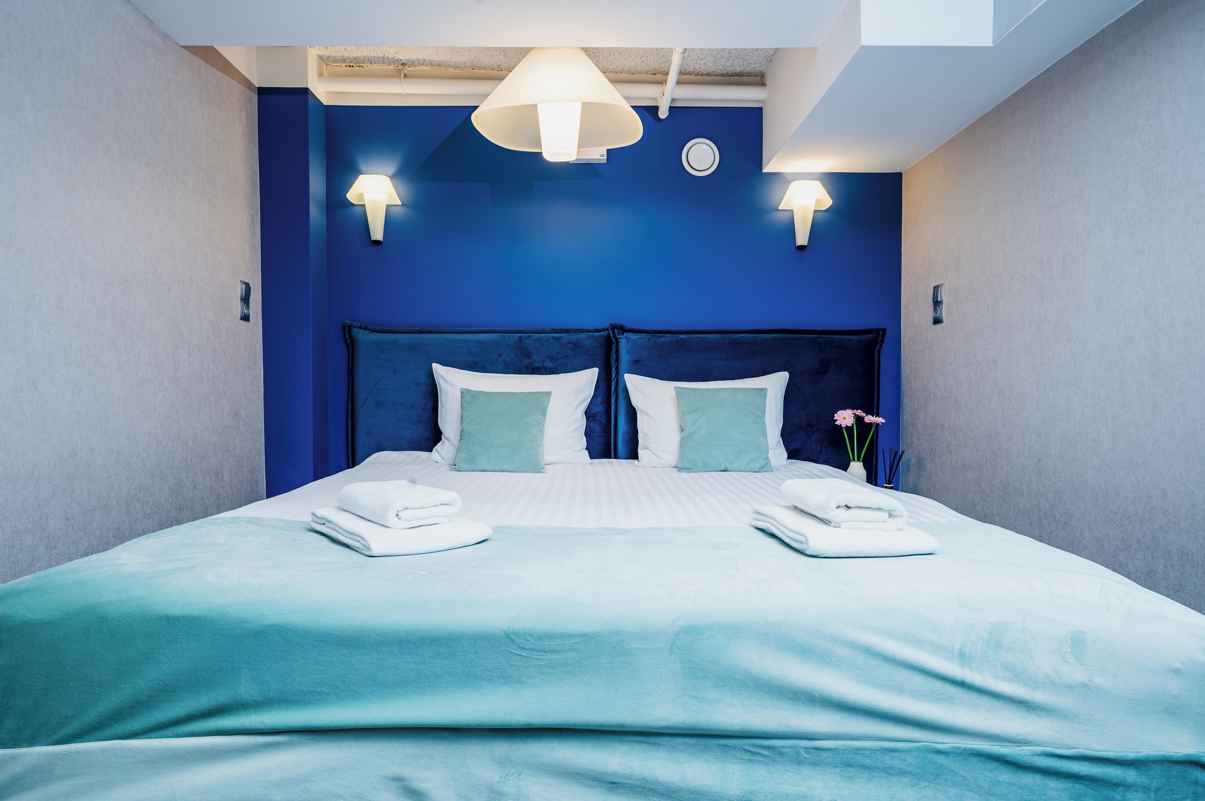 Moderni hotellihuone, jossa on suuri sänky, kaksi yöpöytävalaisinta ja maljakko kukkineen.