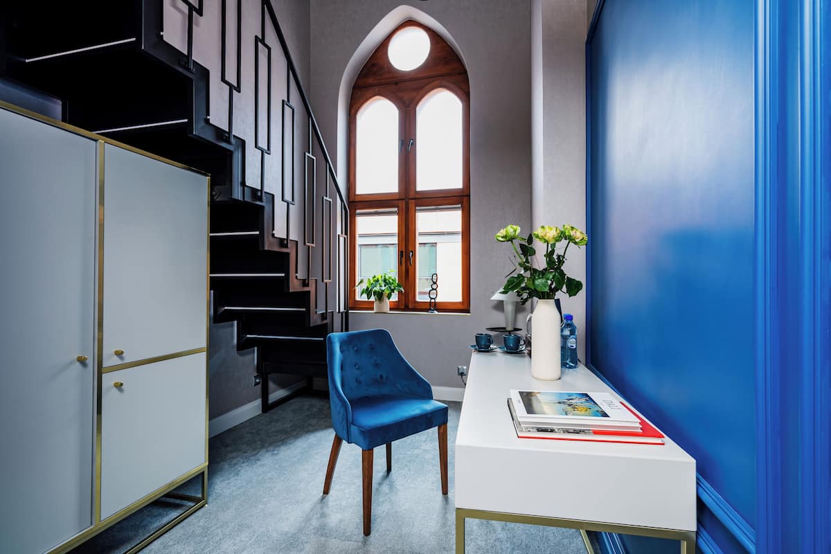 A modern interior with a staircase, a blue velvet chair, a white desk with books, and a vase with flowers.
