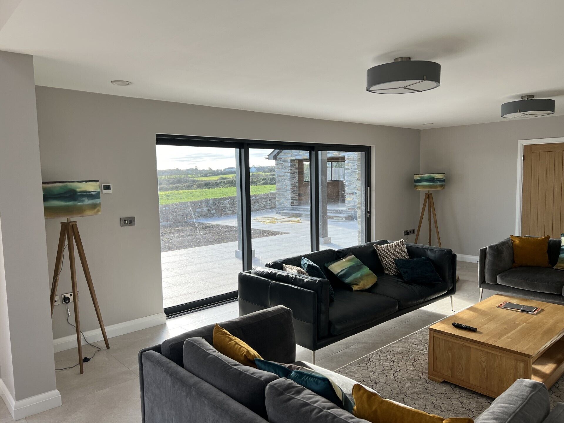 A spacious living room with a large sliding glass door leading to an outdoor area, a black sofa with multiple cushions, a wooden coffee table, and a painting on a tripod stand.