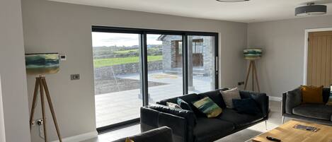 A spacious living room with a large sliding glass door leading to an outdoor area, a black sofa with multiple cushions, a wooden coffee table, and a painting on a tripod stand.