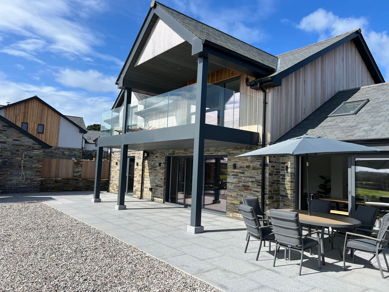 Une maison moderne à deux étages, dotée d’une façade en pierre, d’une véranda en verre et d’un espace extérieur aménagé avec une table et des chaises.