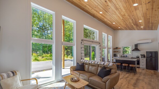 A spacious living room with a leather sofa, a wooden coffee table, and large windows offering a view of the outdoors. - Black Haven Tiny Home (Surry)