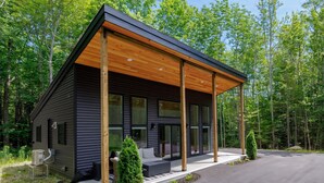 A modern cabin with a covered porch, large windows, and a well-maintained surrounding landscape. - Black Haven Tiny Home (Surry)