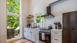 A modern kitchen with white cabinets, a stainless steel stove, and a large window overlooking a forested area. - Black Haven Tiny Home (Surry)