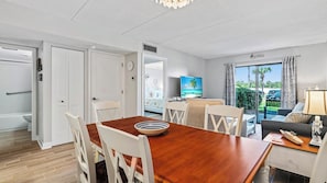A modern hotel room with a wooden dining table, white chairs, a TV, and a view of the outdoors.