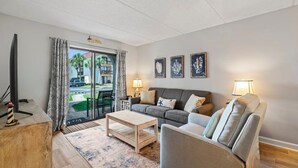 Un salon comprenant un canapé, un fauteuil, une table basse et deux lampes. Il y a une grande fenêtre avec des rideaux, une porte et une vue sur l’extérieur.