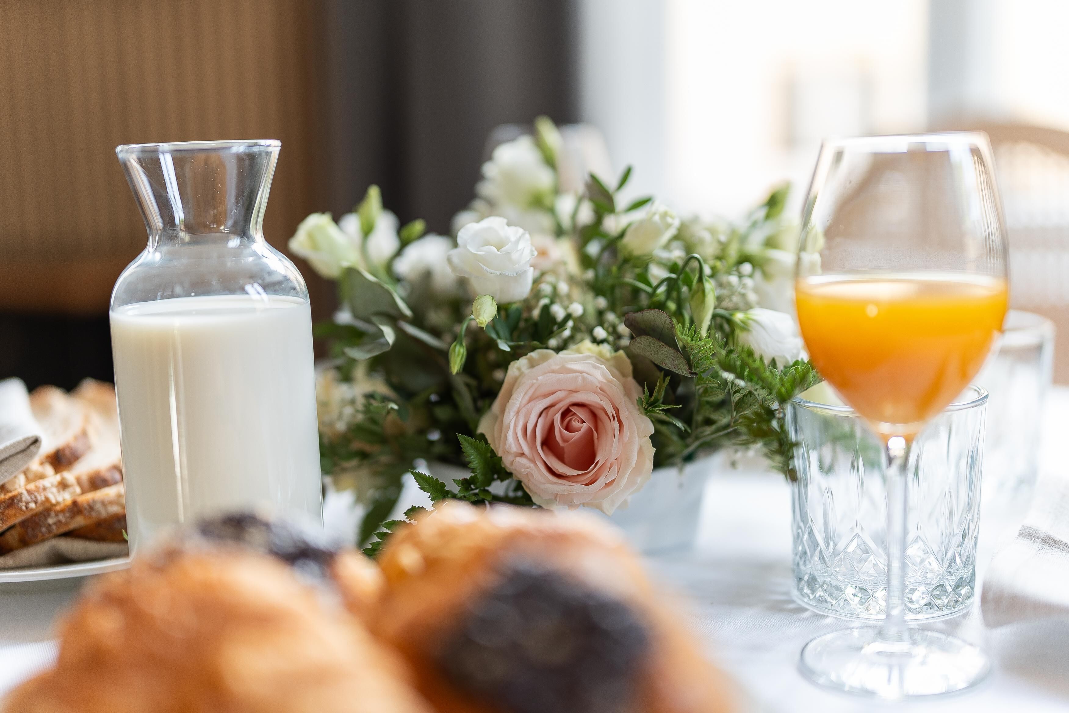 Um cenário de café da manhã com um garrafa de leite, um copo de suco de laranja, doces e um arranjo floral.