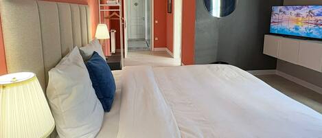 A modern hotel room with a large bed, a TV, a wardrobe, and a bathroom visible through an open door.