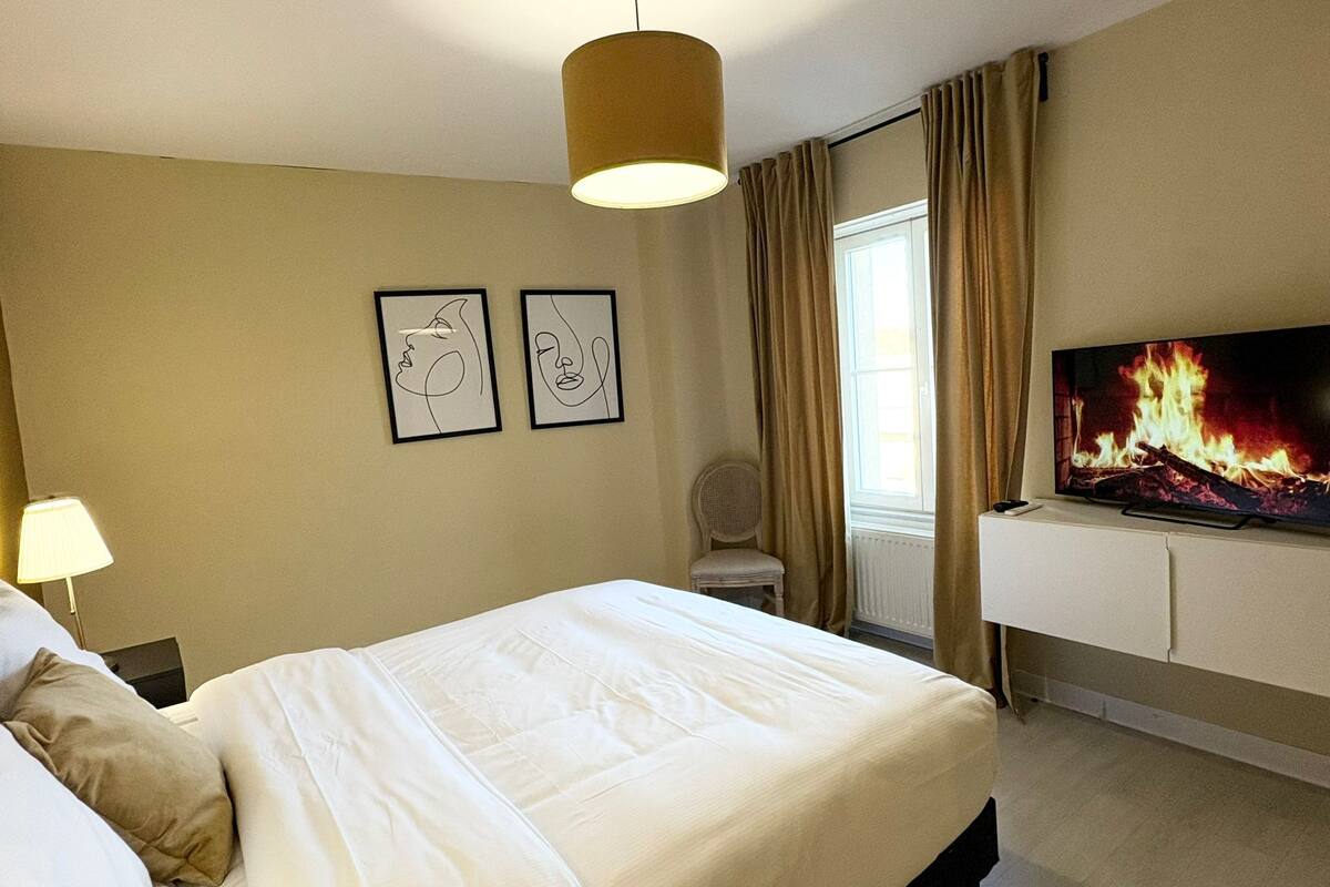 A hotel room with a large bed, a television displaying a fire, two framed artworks on the wall, and a chair near a window.
