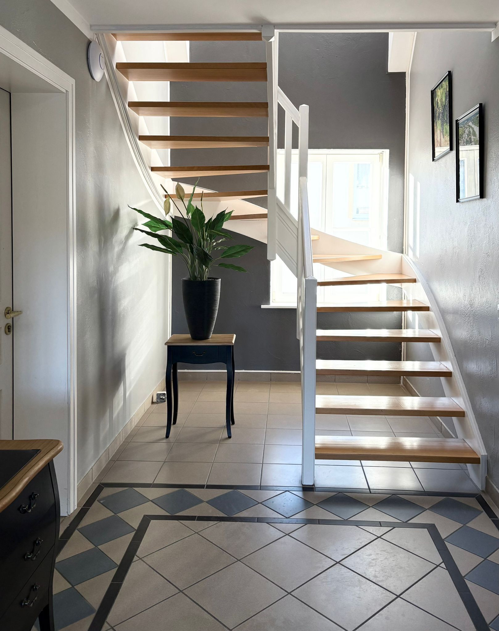 Una escalera con peldaños de madera, barandillas blancas, una planta en maceta y un suelo de baldosas con un diseño geométrico.