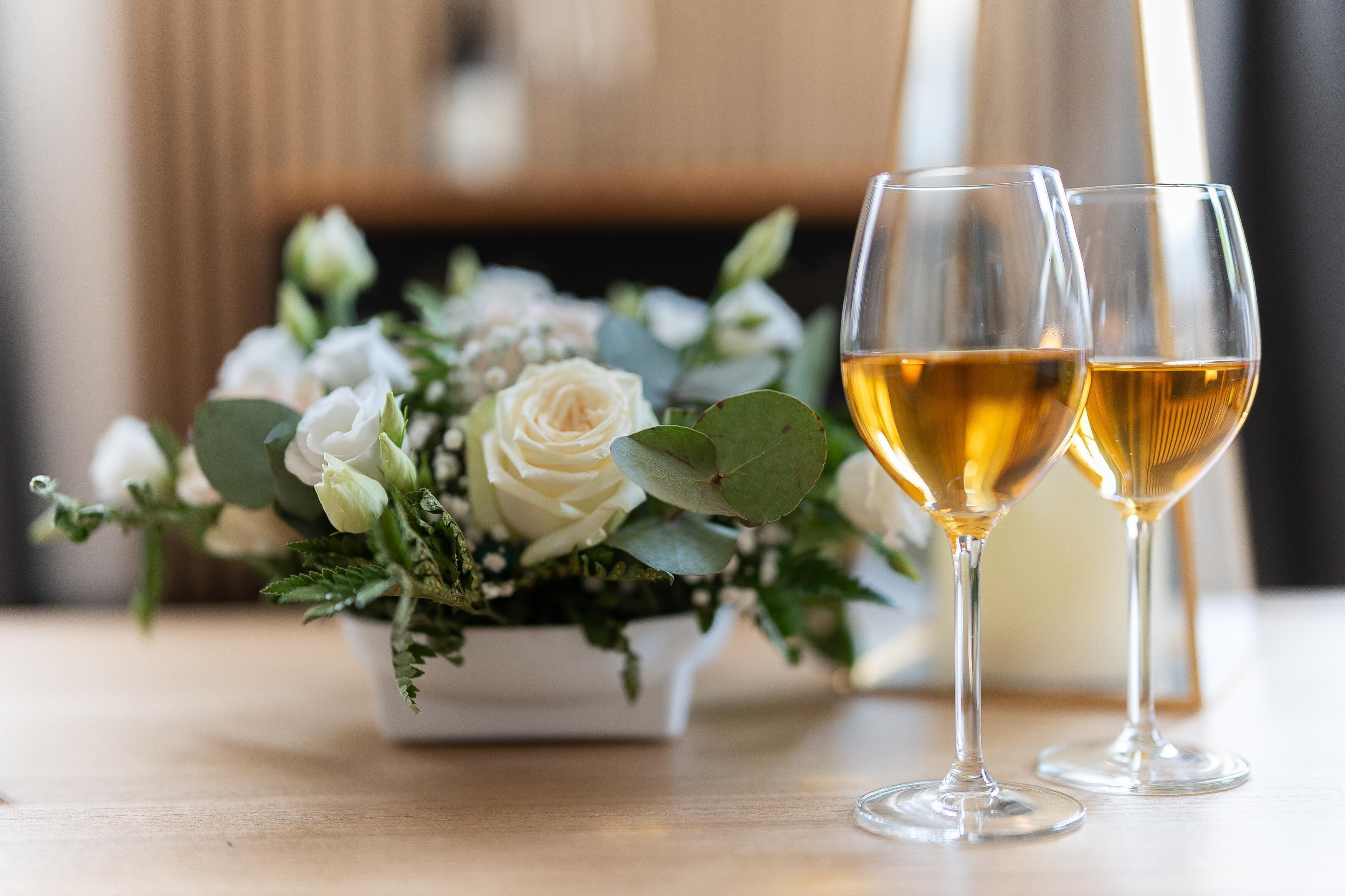 Two glasses of white wine on a wooden table with a floral arrangement.