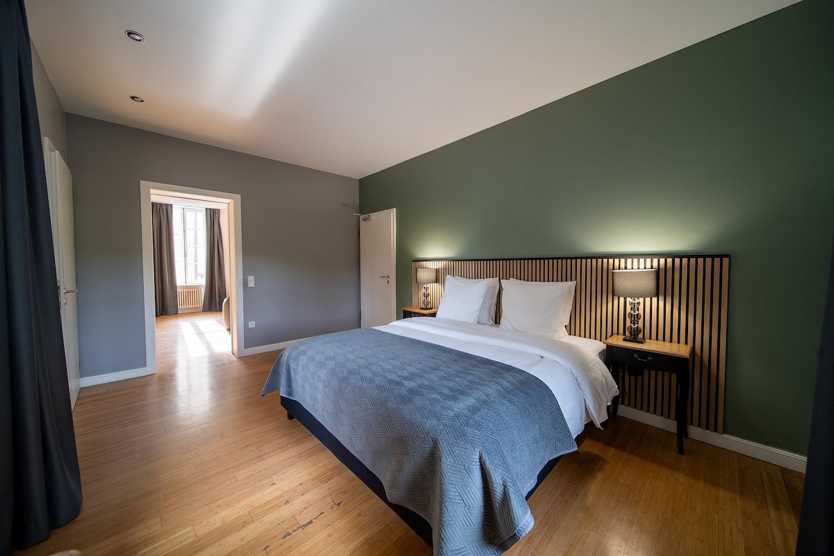 A bedroom with a bed, wooden floor, green walls, and a nightstand with a lamp.