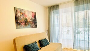 Un salón acogedor con un sofá color beige, dos cojines estampados, un cuadro en la pared y un ventanal con cortinas translúcidas.
