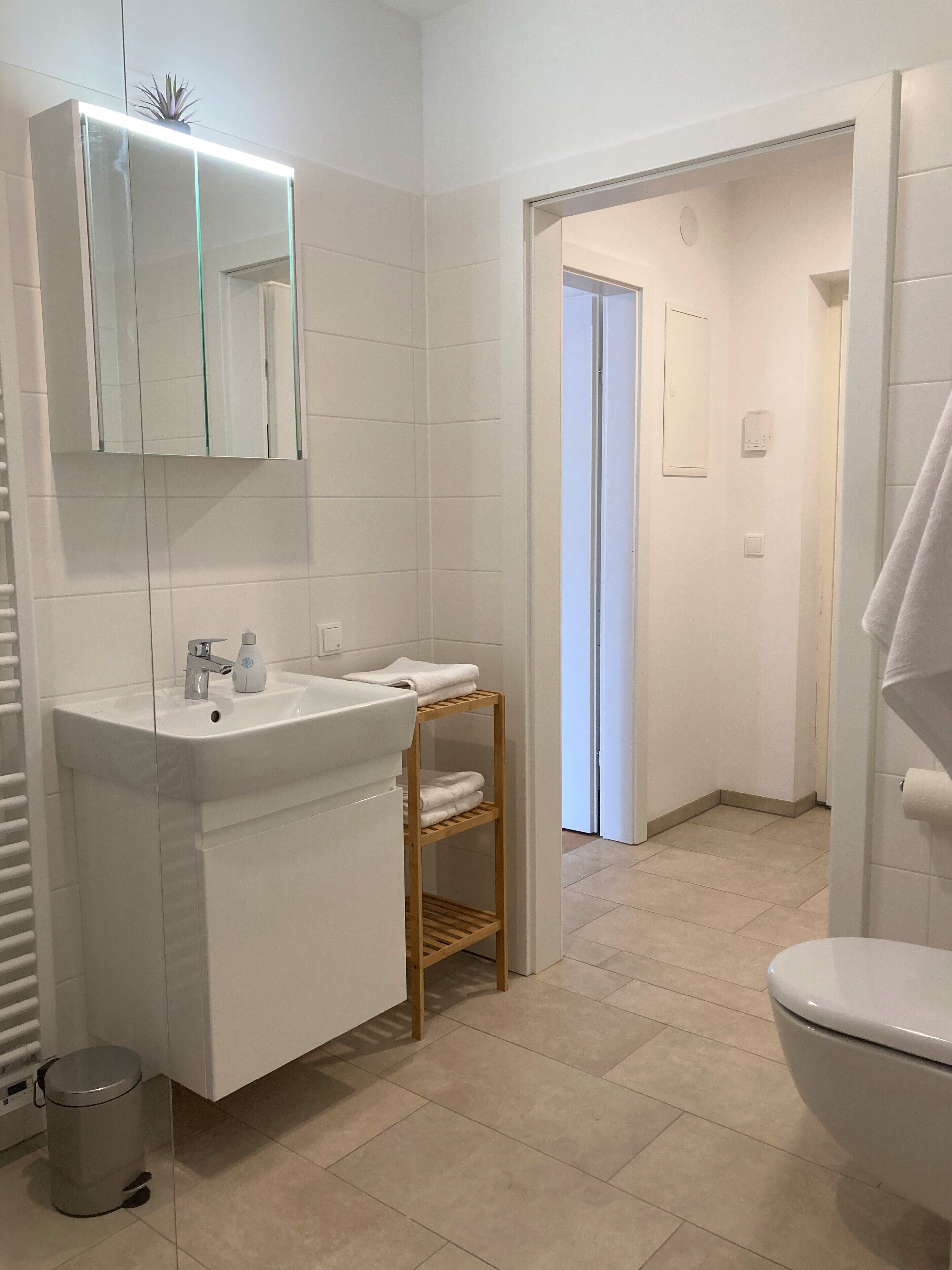 A modern bathroom with a white sink, mirror, and toilet. There is a wooden shelf with towels and a trash can.