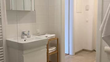 A modern bathroom with a white sink, mirror, and toilet. There is a wooden shelf with towels and a trash can.