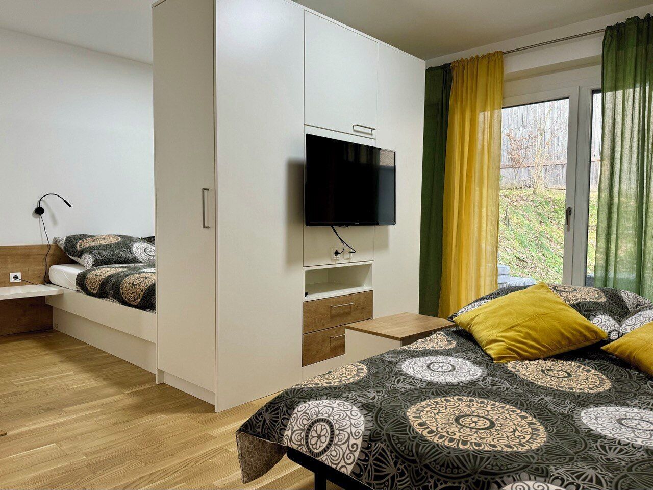 Un dormitorio con una cama, televisor montado en un mueble, mesita de noche y ventana con cortinas.
