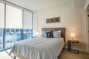 Un dormitorio con un ventanal grande, una cama con ropa de cama a rayas, una mesita de noche con lámpara y un adorno en la pared.
