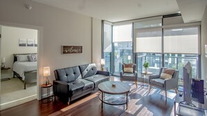A modern living room with a sofa, coffee table, and armchairs. There is a bed with a striped bedspread and a "Welcome" sign on the wall. The room has large windows and a balcony with a view of buildings. - 3380 Peachtree Rd NE Atlanta, GA 30326, USA (Atlanta)