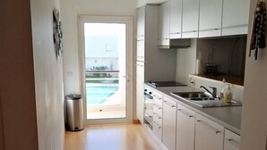 Une cuisine moderne avec des meubles blancs, un évier en acier inoxydable et une vue sur une piscine, visible à travers une porte ouverte.