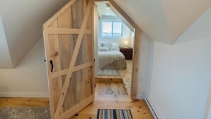 Une chambre à coucher aménagée en mezzanine, avec une porte en bois donnant sur un lit recouvert d’une couvre-lit à motifs floraux.
