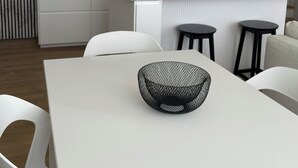 Une cuisine moderne avec des meubles blancs, un luminaire noir suspendu et une table à manger blanche surmontée d’un saladier noir.