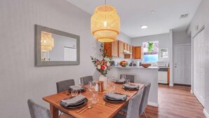 A dining area with a wooden table set for four, a hanging rattan light fixture, and a kitchen in the background. - The Prairie at Brighton | UF • Pool • Workspace (Gainesville)